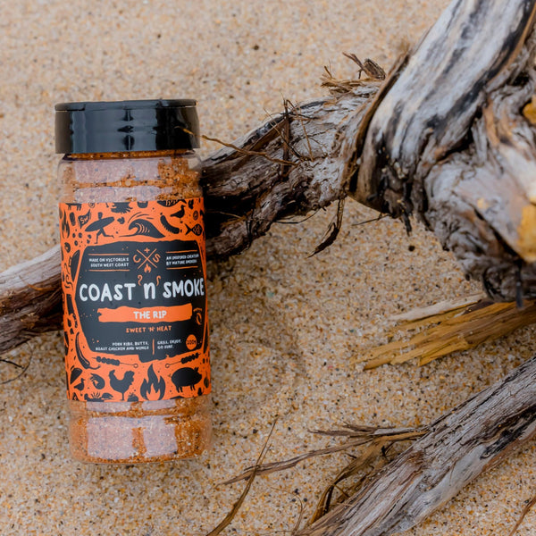 The Rip, a bbq seasoning with a bright orange label, sitting on the beach against drift wood.