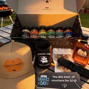 A Coast n Smoke gift box set displayed on a wooden table outdoors, featuring an open box with multiple small bottles, a tan Coast n Smoke cap, a black stubby holder, a bottle of hot sauce, a thermometer, and a Father's Day voucher card.